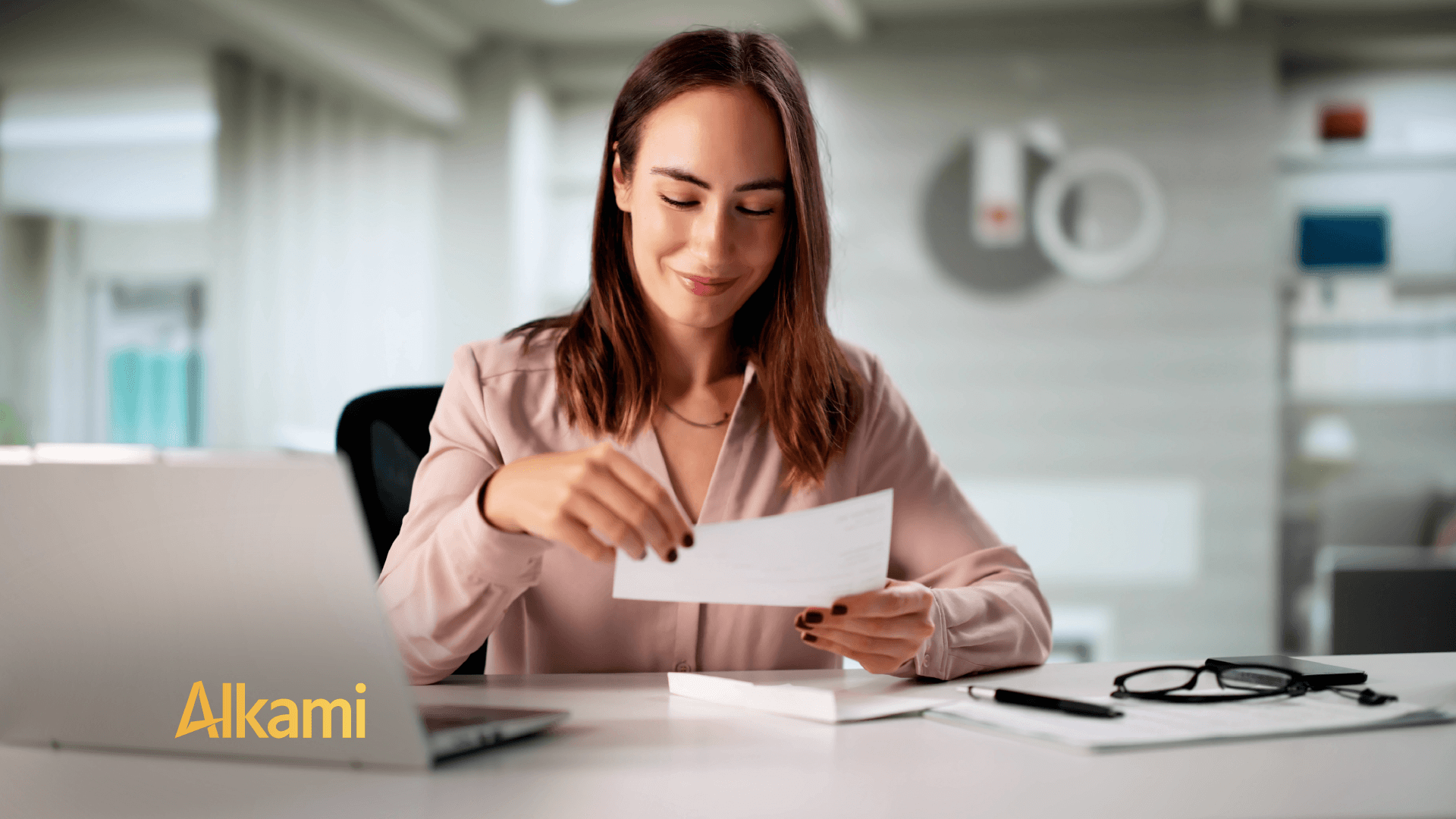 Woman in a pink blouse reviews a document at a desk, smiling, with a laptop and glasses nearby; Alkami logo in the corner.