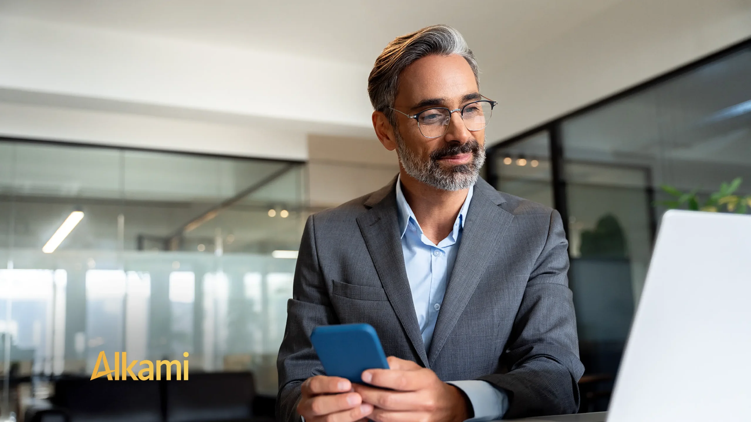 Professional man in a gray suit and glasses sits at a desk in a modern office, looking at a laptop while holding a blue smartphone. Watermark shows 'Alkami' in the corner.