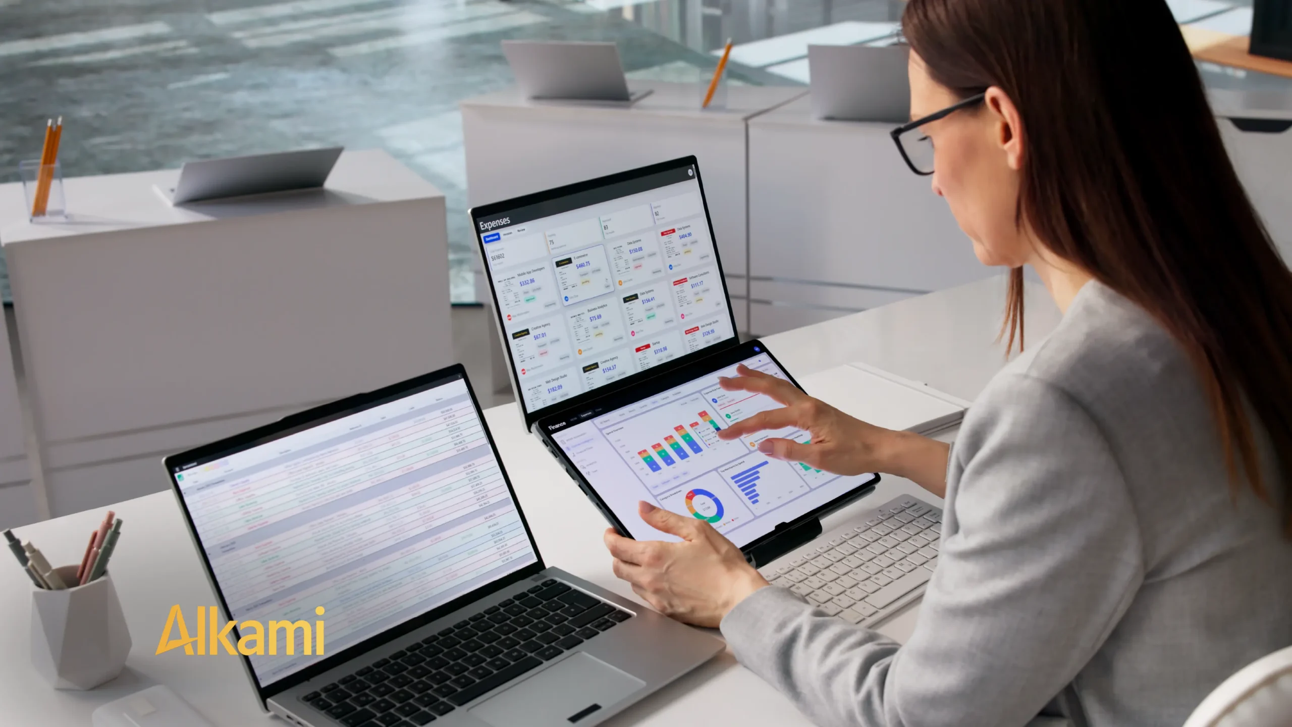Woman wearing glasses uses a laptop and tablet to view financial dashboards at a bright white desk; Alkami logo visible.