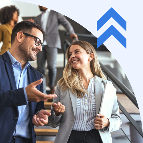 Two coworkers in a modern office stairwell chat and smile, blue logo in the top-right corner, conveying collaboration.