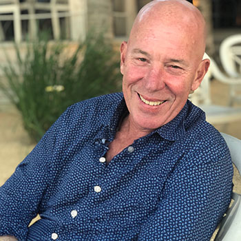 Smiling bald man in a blue patterned shirt sits outdoors near greenery at a cafe.