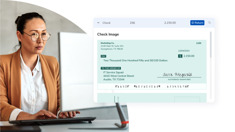 Businesswoman in glasses and a tan blazer typing on a laptop beside a large screen showing a check image.