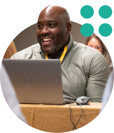 Smiling man using a laptop at a workshop, seated at a table with a yellow lanyard.