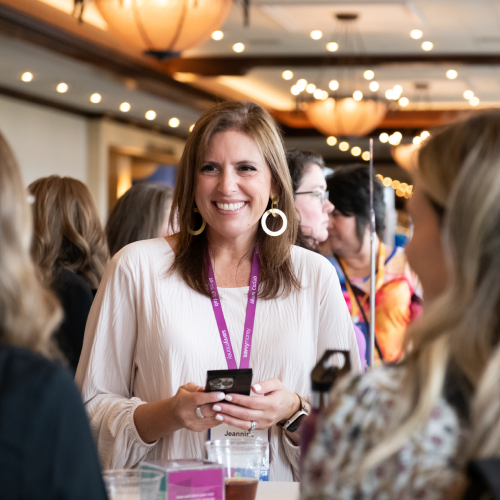 Smiling woman in a white blouse and purple lanyard at a crowded conference, holding a phone at a booth.