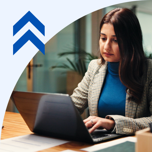 Woman in a blue sweater and blazer typing on a laptop at a desk in a bright office, with an abstract blue chevron logo in the upper left.