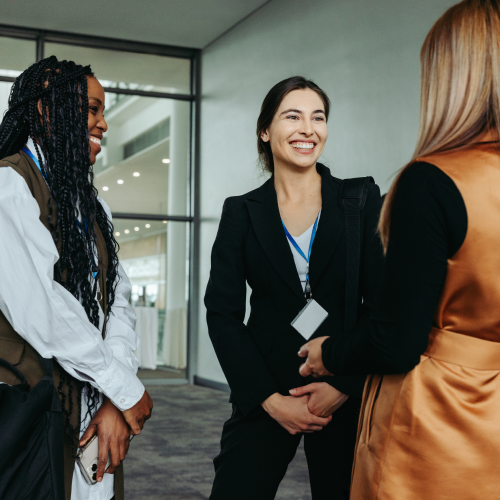 Three women in business attire stand outside a modern building, smiling and talking.