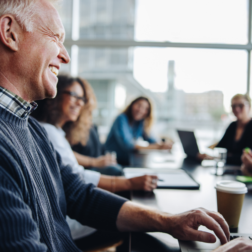 Smiling man in a sweater seated at a conference table with coworkers in a bright meeting room .