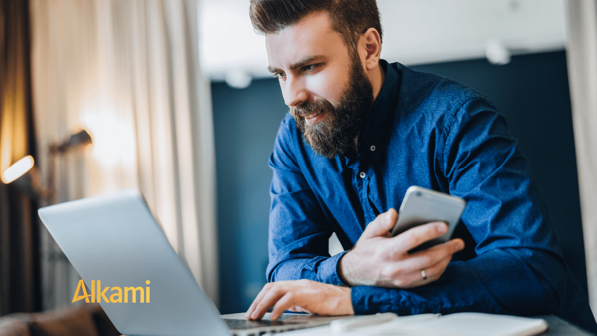 Bearded man in a blue shirt typing on a laptop while holding a smartphone in a bright workspace; Alkami logo visible in the corner.