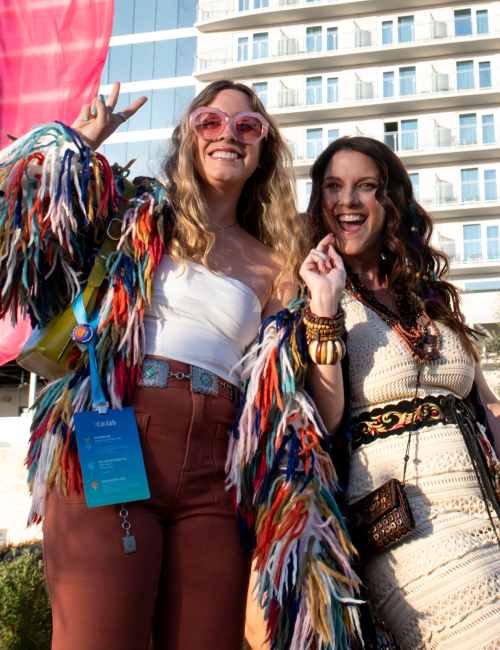 Two friends posing for a sunny outdoor festival photo; one wears a colorful fringe jacket and pink sunglasses, the other in a cream crochet dress with jewelry.