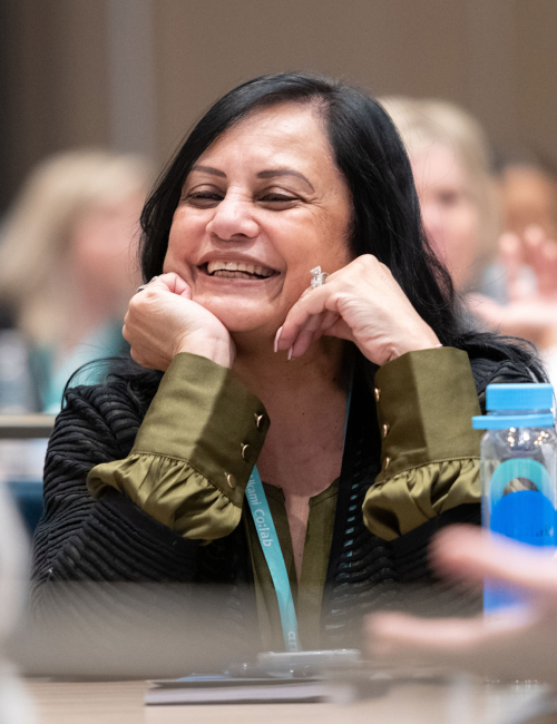 Smiling woman with black hair resting her chin on her hands at a conference table, wearing a green blouse and olive jacket with a lanyard visible.