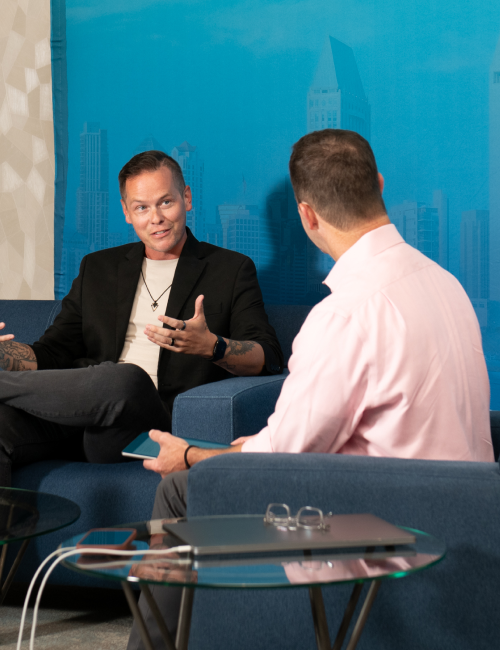 Two men in a modern office lounge converse on a blue couch, one gesturing while the other listens.