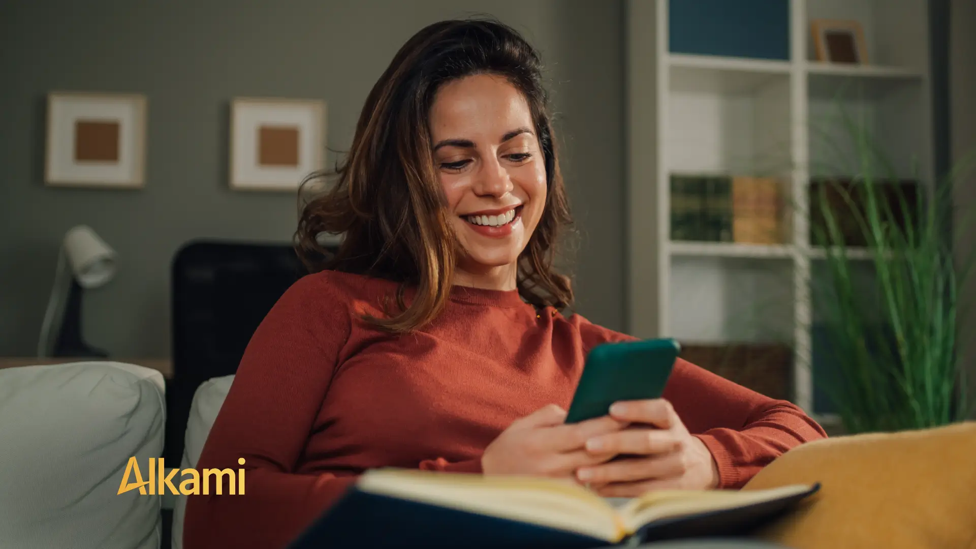 Woman relaxing on a sofa, smiling at her green smartphone, with an open notebook nearby; Alkami logo bottom left.