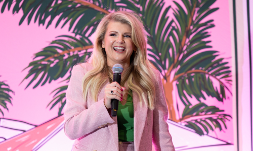 Woman on stage holding a microphone, smiling, wearing a pink blazer against a pink, palm-leaf backdrop.