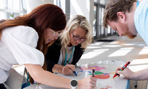 Three people—two women and a man—lean over a table, drawing with markers on a colorful poster in a bright space.