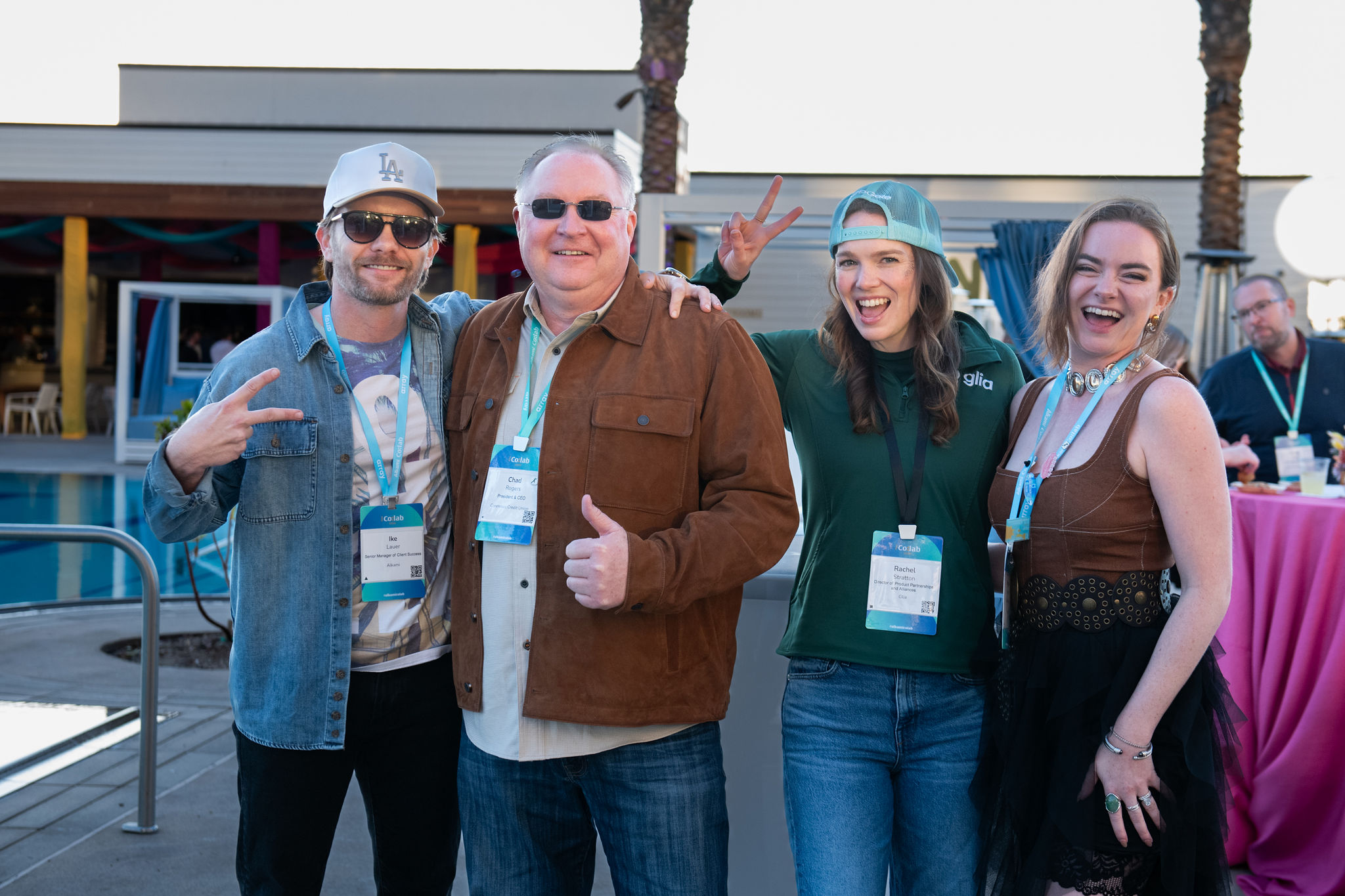 Group of four adults smiling at camera at an outdoor event, wearing lanyards and casual clothing by a poolside area.