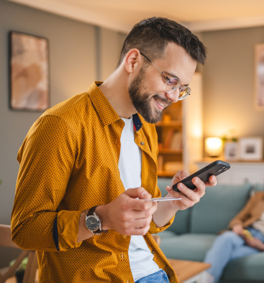 Man wearing a mustard shirt and glasses smiles while holding a credit card in one hand and a smartphone in the other in a cozy living room.